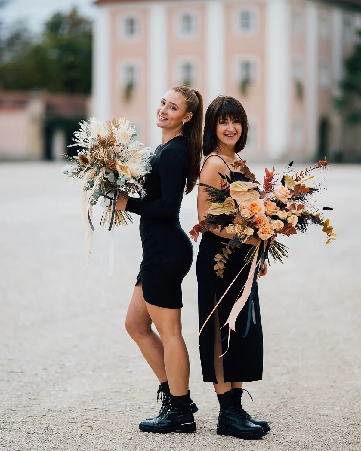 Zwei lächelnde Frauen in schwarzen Kleidern stehen Rücken an Rücken und halten große Blumensträuße, im Hintergrund ein historisches Gebäude