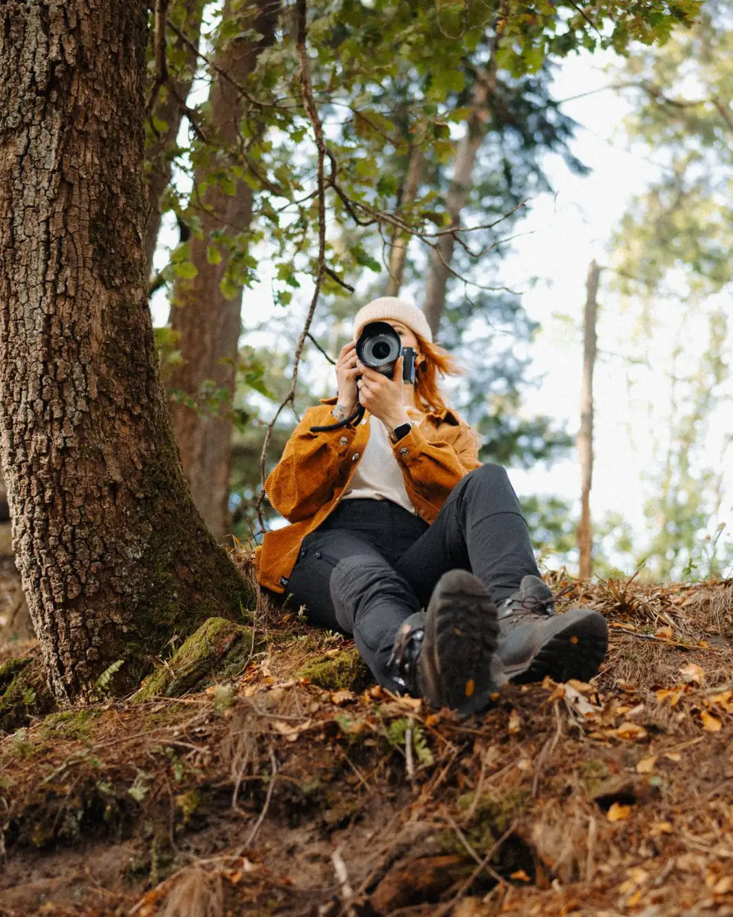 Fotografin mit heller Mütze und ockerfarbener Jacke sitzt an einem Baum im Wald und fotografiert in Richtung Kamera