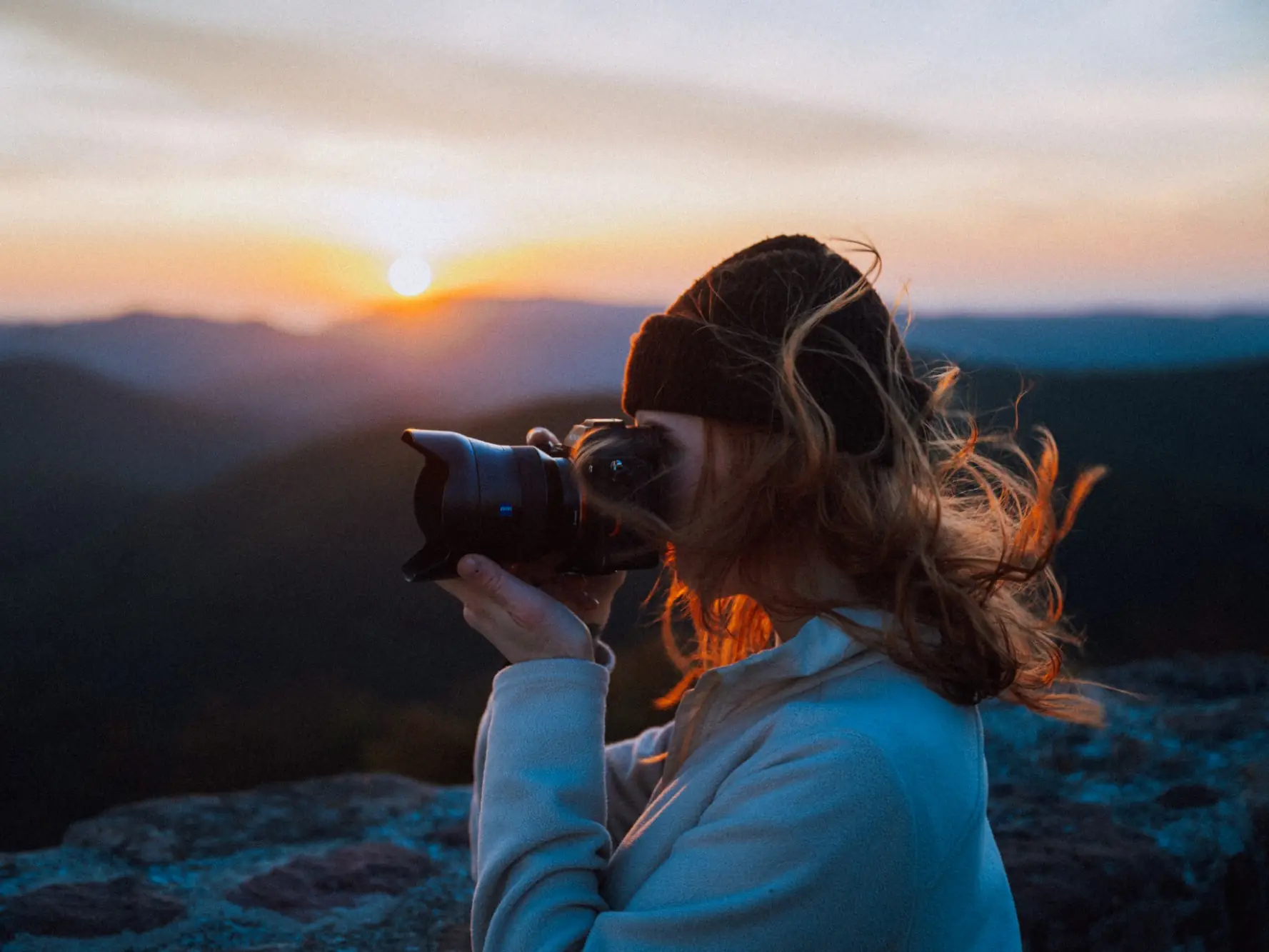 Fotografin mit Mütze und wehendem Haar fotografiert bei Sonnenaufgang auf einem Felsen über bewaldeten Hügeln