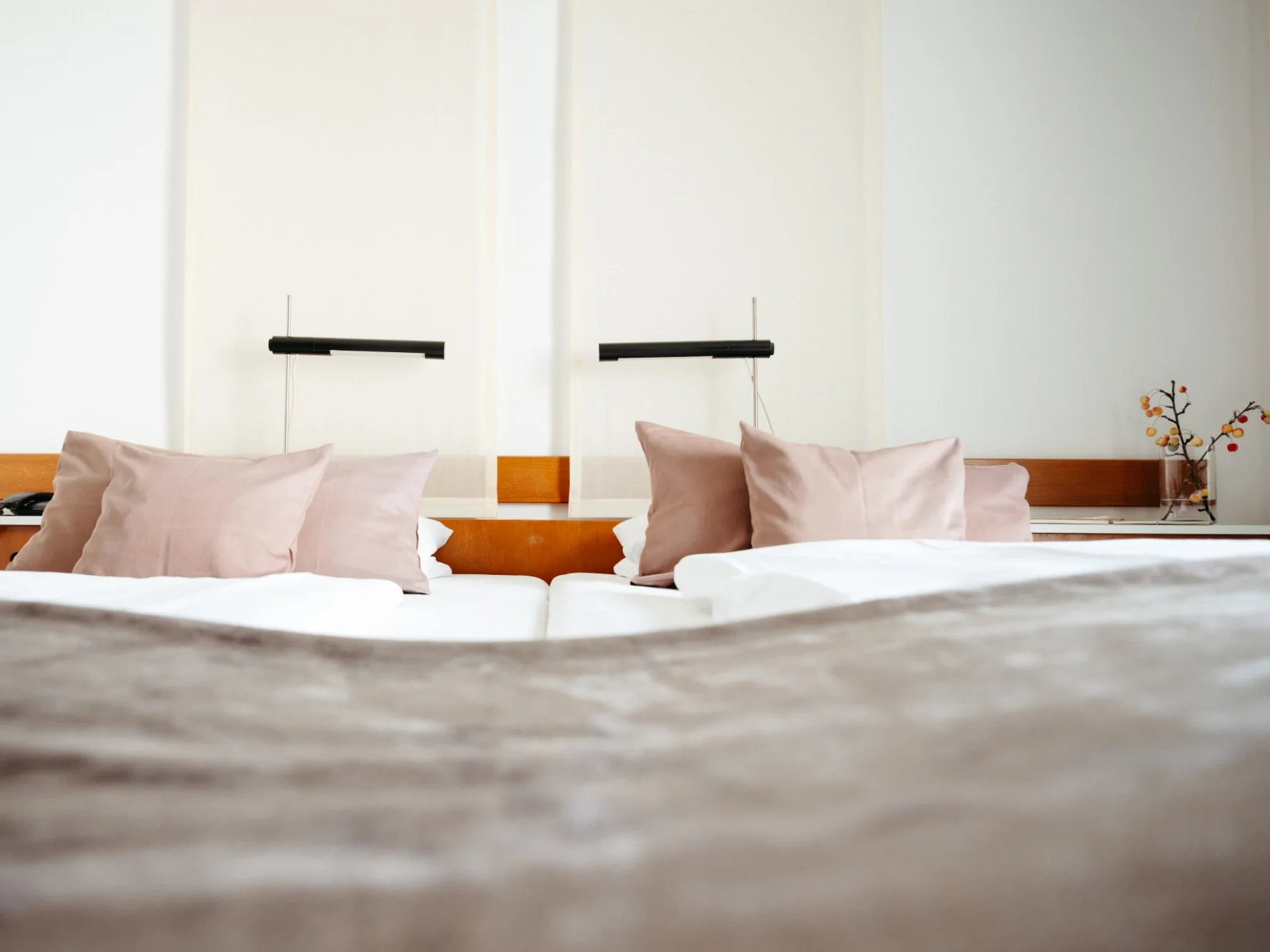 Hotelzimmer mit Doppelbett, rosafarbenen Kissen, zwei schwarzen Leseleuchten und einem Zweig in einer Glasvase