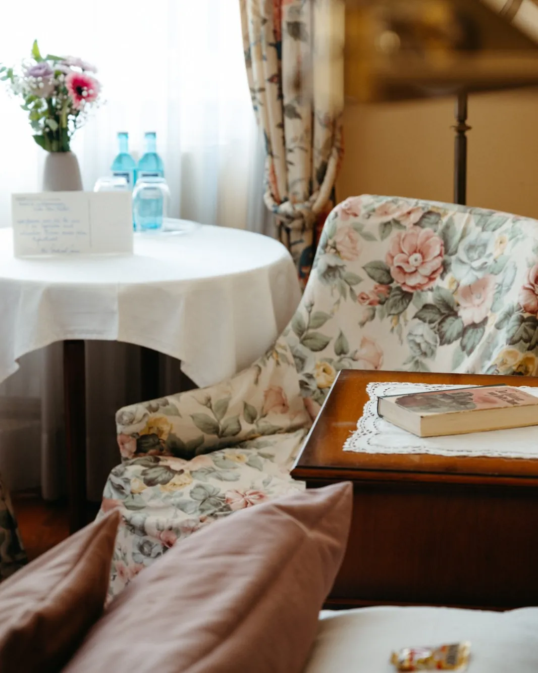 Hotelzimmer mit geblümtem Sessel, rundem Tisch mit Blumen und Wasserflaschen und einem Buch auf dem Nachttisch