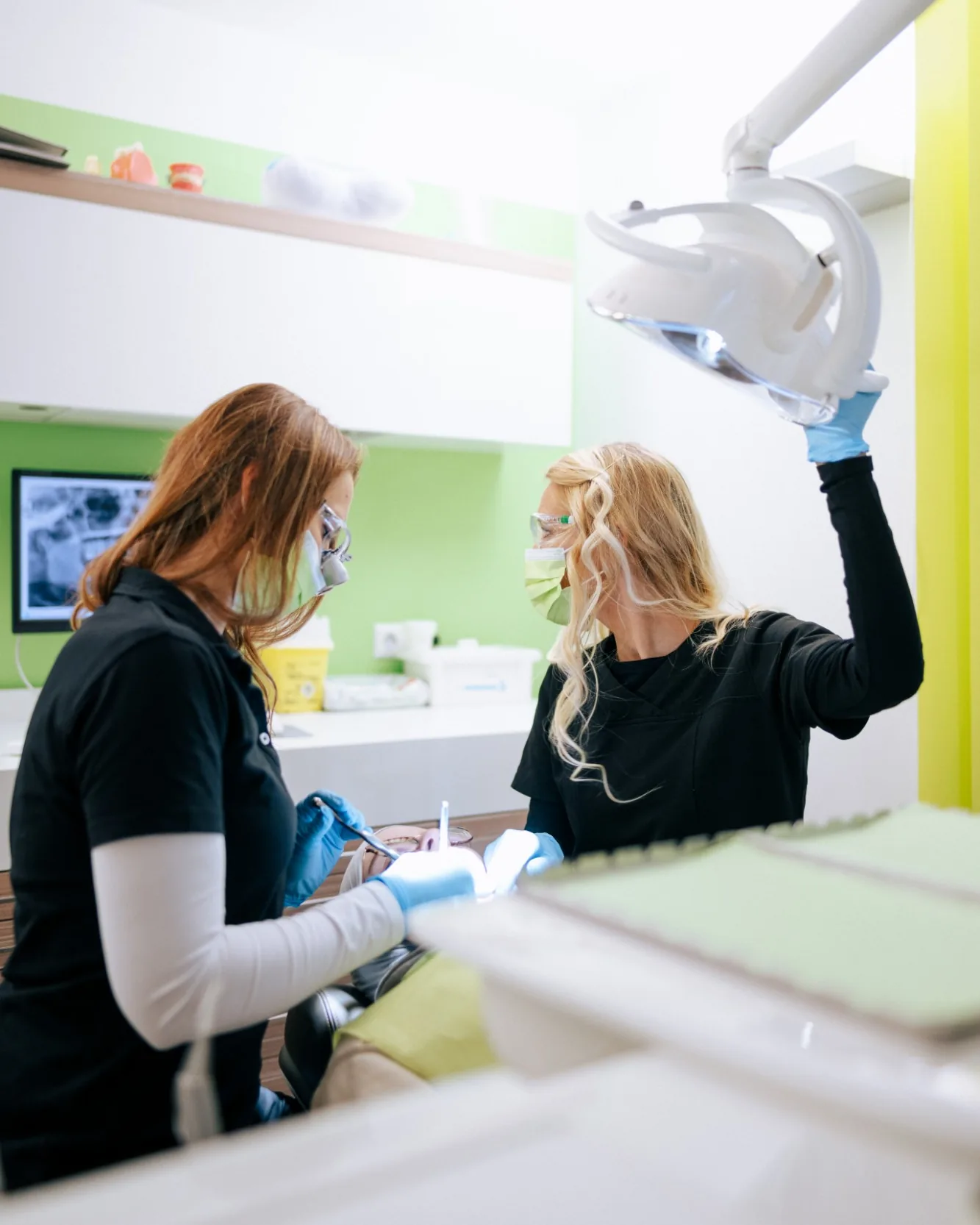 Zwei Mitarbeiterinnen einer Zahnarztpraxis mit Mundschutz und Schutzbrille bei einer Behandlung, eine richtet die Behandlungsleuchte aus