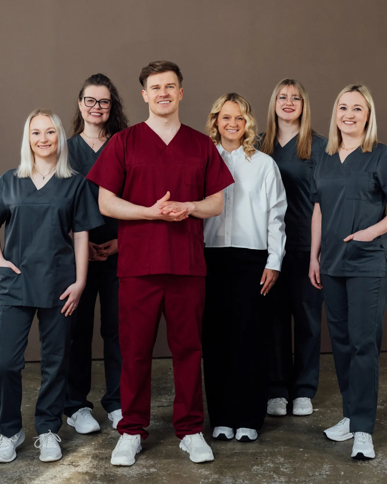 Sechsköpfiges Praxisteam in Arbeitskleidung steht lächelnd dicht beieinander vor einer braunen Wand