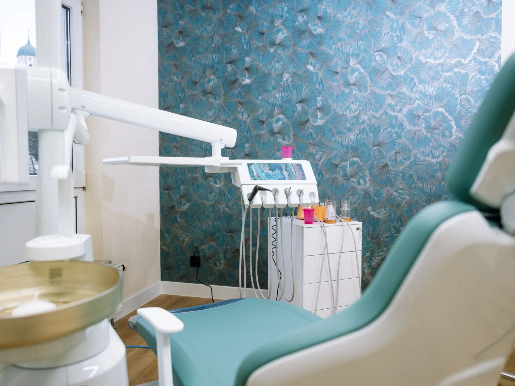 Behandlungszimmer einer Zahnarztpraxis mit türkisfarbenem Behandlungsstuhl, Instrumenteneinheit und blau gemusterter Tapete, Blick über die Speischale