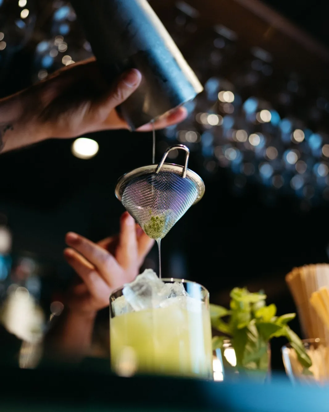 Ein Cocktail wird an der Bar durch ein feines Sieb in ein Glas mit Eis gegossen, daneben frische Minze