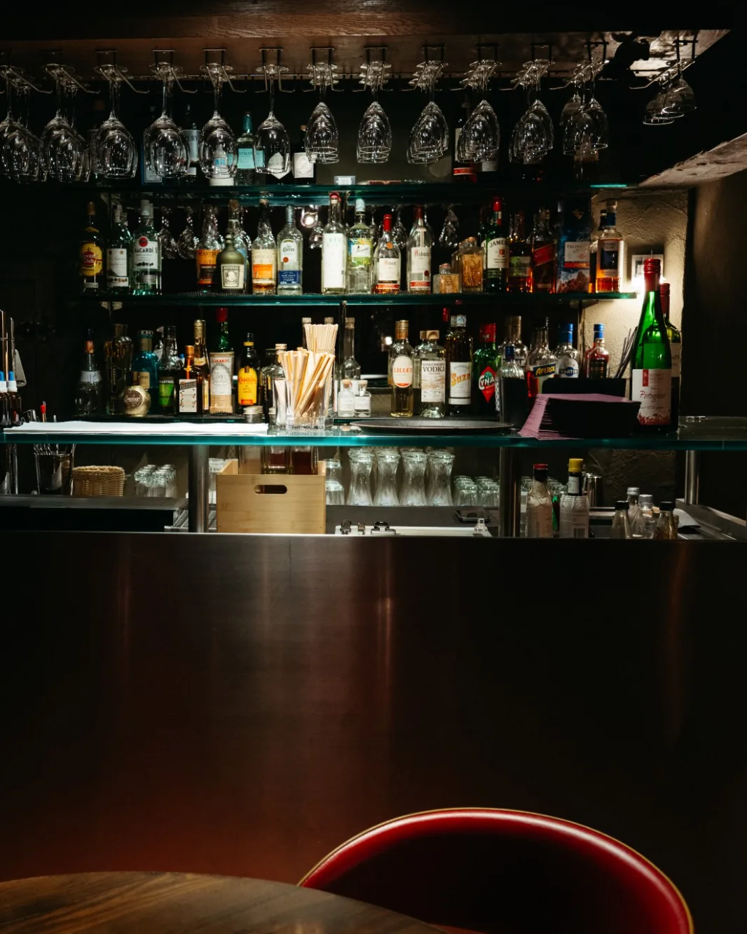 Beleuchtete Bar eines Restaurants mit Flaschenregal, hängenden Weingläsern und einem roten Stuhl im Vordergrund