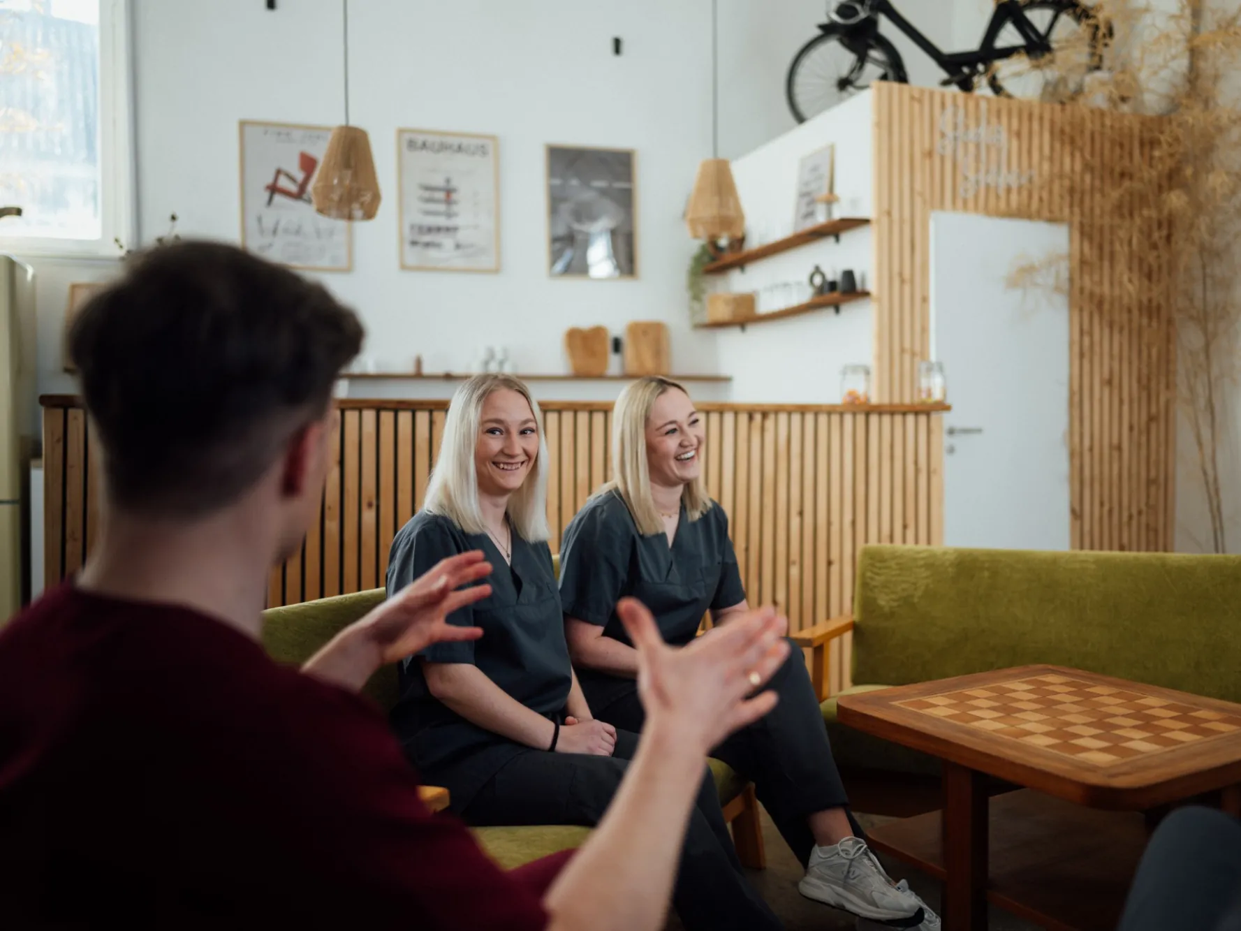 Zwei lachende Mitarbeiterinnen in dunkler Praxiskleidung sitzen auf einem grünen Sofa und hören einem gestikulierenden Kollegen im Vordergrund zu