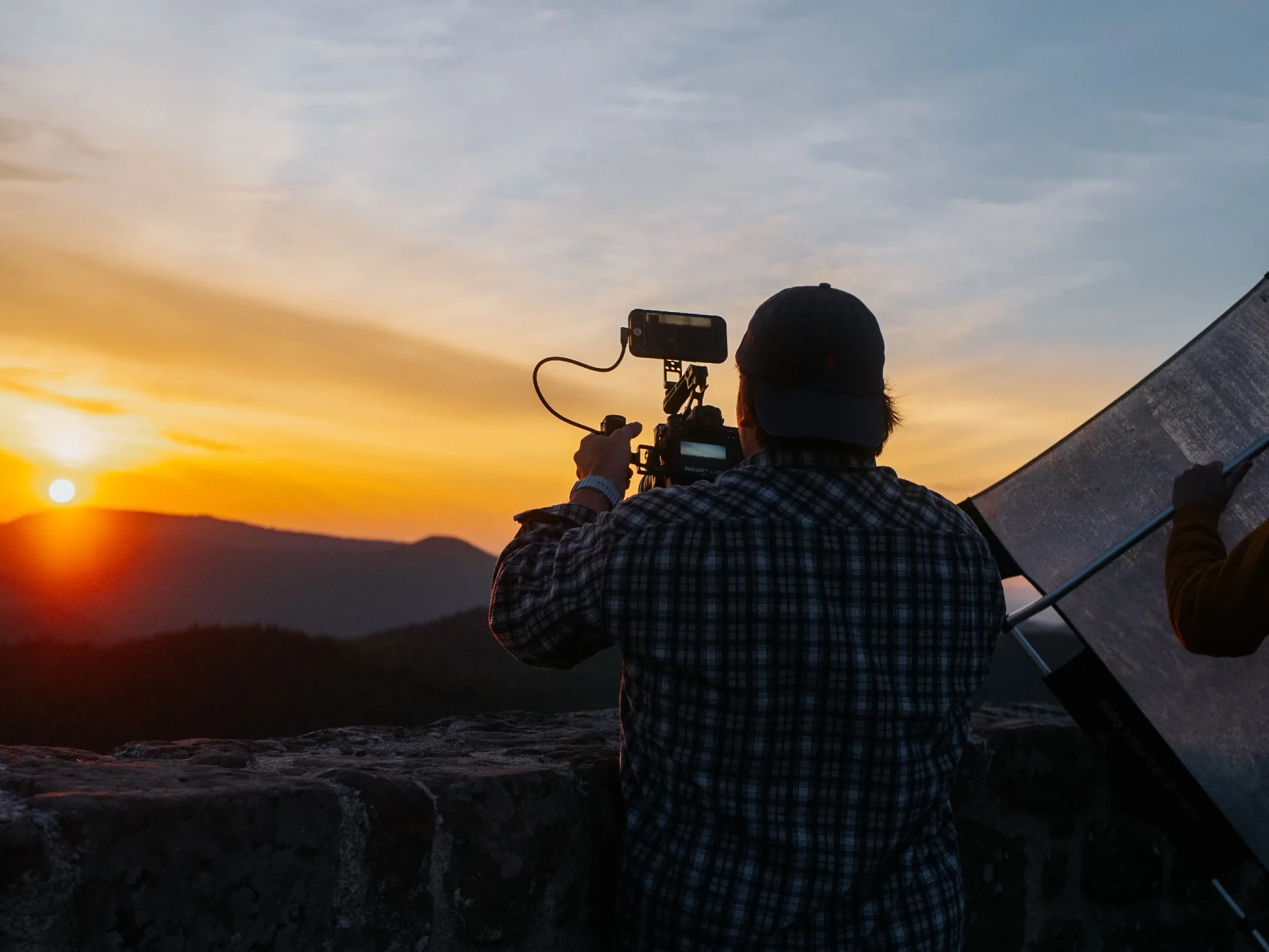 Nick Paysan filmt von hinten gesehen mit einer Videokamera den Sonnenaufgang über bewaldeten Bergen